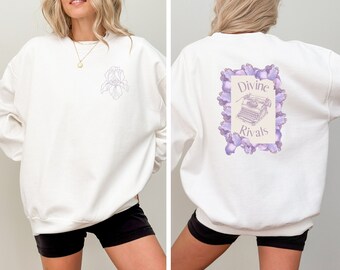 May include: White sweatshirt with a floral design on the front and back. The front features a light purple iris. The back displays the text "Divine Rivals" with a typewriter illustration, framed by purple flowers.