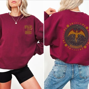 May include: Burgundy sweatshirt with "FLY OR DIE" text on the front and a circular graphic on the back featuring a dragon, clouds, and the word "BASGARTH". The design is in gold and brown tones.