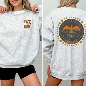 May include: Light gray sweatshirt with the text "FLY OR DIE" on the front and a circular graphic on the back featuring a phoenix, clouds, and the word "BASGIATH". The design includes gold and brown accents.