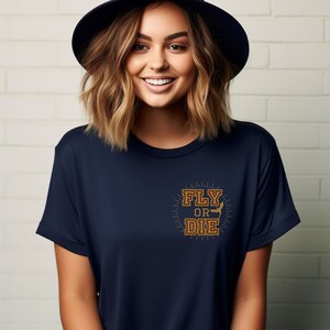 May include: Navy blue t-shirt with the text "FLY OR DIE" in a gold, vintage-style design. The graphic includes a silhouette of a bat. The shirt is a short-sleeved crew neck.
