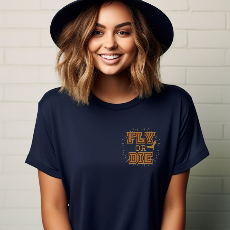 May include: Navy blue t-shirt with the text "FLY OR DIE" in a gold, vintage-style design. The graphic includes a silhouette of a bat. The shirt is a short-sleeved crew neck.