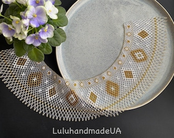 Puede incluir: Un collar de cuentas hecho a mano con un patrón geométrico en blanco, dorado y marrón. El collar se exhibe en un plato azul claro con un arreglo floral de flores moradas y blancas. El texto "LuluhandmadeUA" es visible.