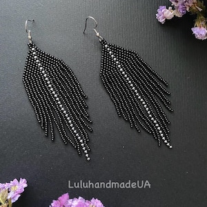 May include: A pair of black beaded earrings with a feather-like design. Each earring features a central line of white beads and dangling strands of black beads. The earrings are displayed on a dark surface with purple flowers.
