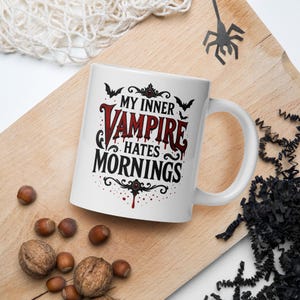 Peut inclure: Mug en céramique blanche avec l'inscription "MY INNER VAMPIRE hates MORNINGS" en noir et rouge. La tasse a une anse et est décorée de chauves-souris et de gouttes de sang. La tasse est sur une surface en bois avec des noix et une décoration d'araignée.