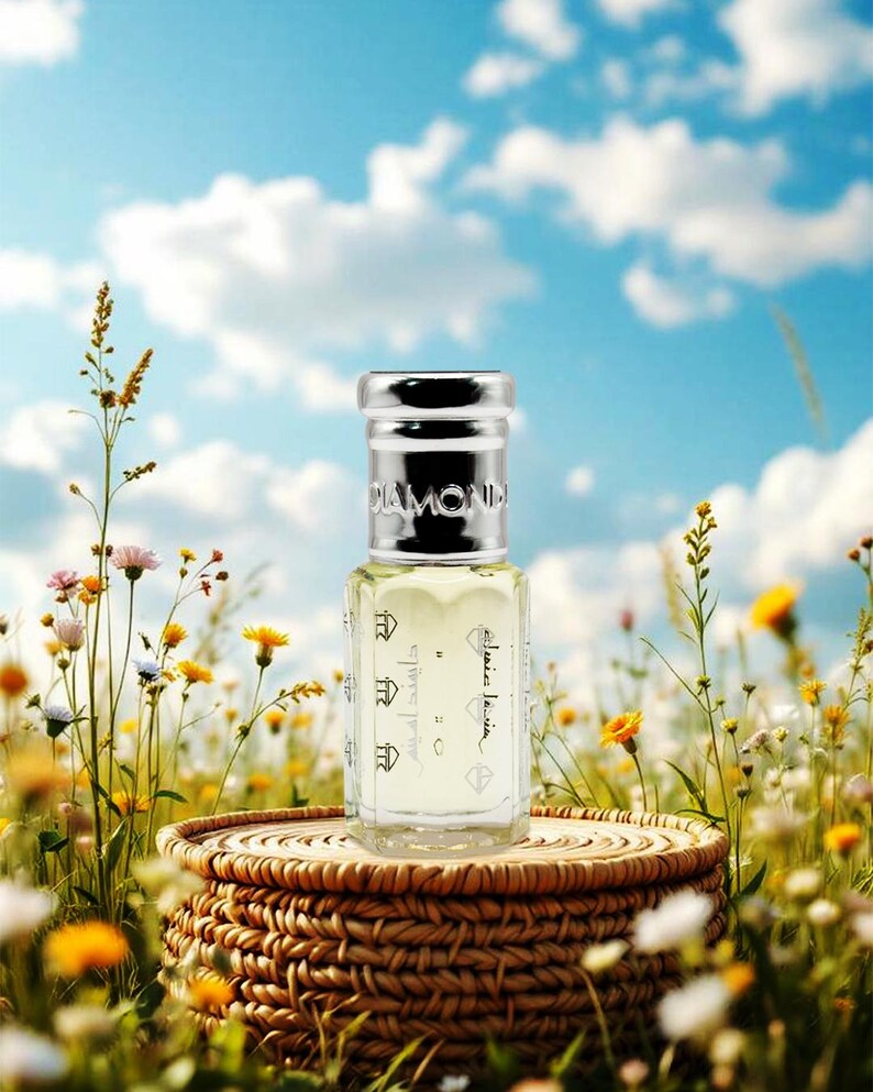 May include: A clear perfume bottle with a silver cap, labeled "DIAMOND", sits on a woven basket amidst wildflowers. The bottle contains a light yellow liquid and has Arabic text. The background features a blue sky with fluffy white clouds.