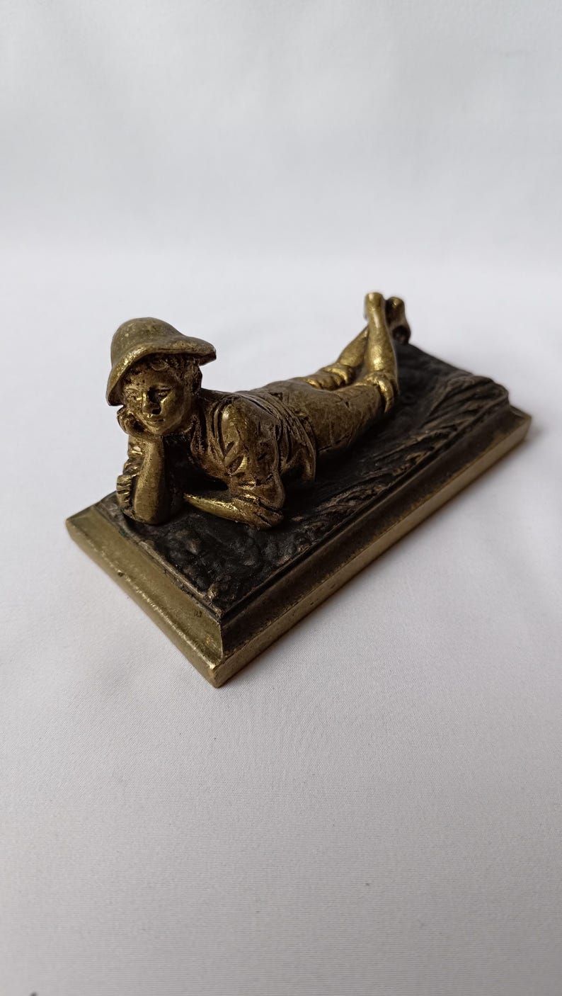 May include: A bronze figurine of a person reclining on a rectangular base. The figure is wearing a hat and appears to be resting their head on their hand. The base has a textured surface and a raised border.