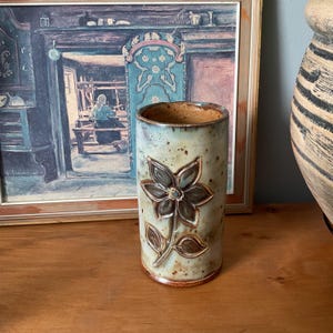 Könnte beinhalten: Eine zylindrische Keramikvase mit floralem Design. Die Vase ist hellblau mit braunen Akzenten und einem erhabenen Blumenmotiv. Der Innenraum ist dunkler braun. Die Vase ist etwa 15 cm hoch.