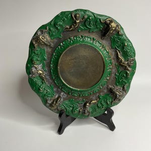 Könnte beinhalten: Ein antiker dekorativer Teller mit grün-goldener Farbgebung. Der Teller hat ein zentrales kreisförmiges Design und ist mit verzierten Relief-Figuren und einem dekorativen Rand verziert. Er wird auf einem schwarzen Ständer präsentiert.