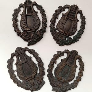 Può includere: Quattro antichi emblemi decorativi color bronzo. Ogni emblema presenta una lira all'interno di una ghirlanda floreale. La lira è uno strumento a corde e la ghirlanda è composta da foglie e fiori. Gli emblemi hanno un aspetto invecchiato.