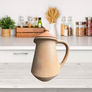Puede incluir: Una jarra de terracota con tapa, de color marrón claro y forma redondeada. La jarra tiene un asa en el lateral y está sobre una superficie de madera.