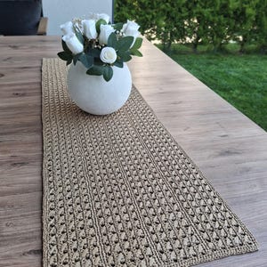 Peut inclure: Un chemin de table rectangulaire en crochet beige avec un motif texturé répétitif. Un vase blanc rond est posé sur le chemin de table, contenant un bouquet de roses artificielles blanches avec des feuilles vertes et des petites fleurs.