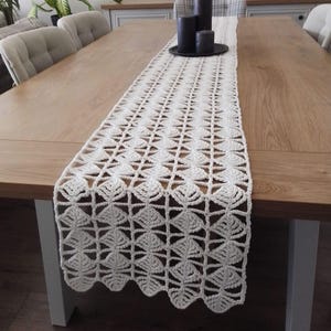 Pode incluir: Um corredor de mesa de crochê branco com um padrão delicado e intrincado. O corredor está colocado sobre uma mesa de madeira com duas cadeiras de cada lado.
