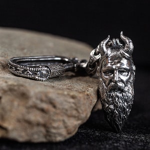 Puede incluir: Un colgante de plata detallado que representa una cara barbuda con cuernos, conectado a un cierre decorativo. El colgante presenta detalles intrincados, con un enfoque en los rasgos faciales y el diseño del cierre. El fondo oscuro resalta el colgante de plata.