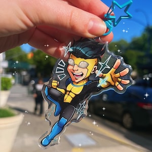 Puede incluir: Un llavero de plástico transparente con un superhéroe de dibujos animados con ropa amarilla y azul. El superhéroe lleva gafas y tiene una expresión decidida. El llavero está decorado con purpurina y estrellas.