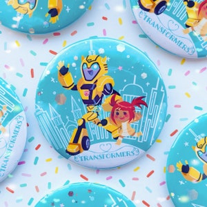 Könnte beinhalten: Ein blauer und gelber Knopf mit einem Cartoon-Bild von Bumblebee von Transformers und einer kleinen menschlichen Figur. Der Knopf trägt den Text "Transformers".
