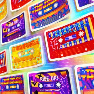 Pode incluir: Uma coleção de adesivos de fita cassete coloridos com vários lemas, incluindo "Rise Up! Peace Through Tyranny", "Til All Are One", "Transform and Roll Out! We Can Do It!" e "The Touch of the Power".