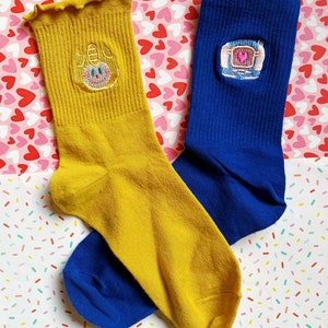 May include: A pair of socks with embroidered designs. One sock is yellow with a cartoon face and the other is blue with a heart-shaped design.