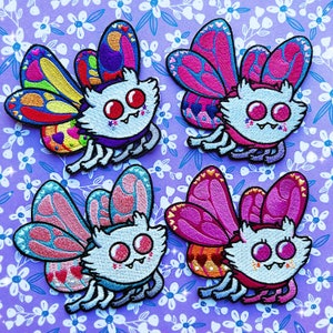 Może przedstawiać: Cztery kolorowe haftowane łatki do prasowania przedstawiające kreskówkowe postacie potworów z skrzydłami. Każda łatka ma inny schemat kolorów i wzór, z akcentami różowego, niebieskiego, czerwonego i żółtego.