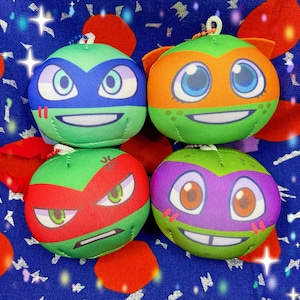 May include: Four plush toys shaped like Teenage Mutant Ninja Turtles. Each turtle has a different coloured mask: blue, orange, red, and purple. The turtles have different facial expressions.