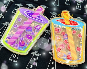 May include: Two clear plastic bubble tea charms with glitter and colorful designs. The charms are shaped like cups with straws and feature a variety of small, colorful images inside. One charm is purple and pink with a pink and white striped straw, while the other is yellow and orange with an orange and yellow striped straw.