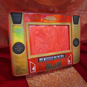 May include: A red and gold retro-style clutch with a clear window. The clutch features black speaker details, a REC button, and play/pause controls. The design resembles an old-fashioned audio player.