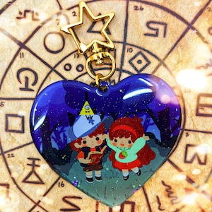 Könnte beinhalten: Ein herzförmiger Schlüsselanhänger mit glitzerndem blauem Hintergrund, auf dem zwei Comicfiguren, ein Junge und ein Mädchen, in einem Wald stehen. Der Junge trägt einen rot-weißen Hut mit einem gelben Dreieck oben drauf, und das Mädchen trägt einen rot-weißen Hut mit einer roten Schleife. Der Schlüsselanhänger ist an einem goldenen sternförmigen Anhänger befestigt.