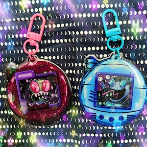 May include: Two colourful plastic keychains shaped like a Tamagotchi toy. The keychain on the left is red and features a green alien with red eyes. The keychain on the right is blue and features a white alien with a purple head and black eyes.