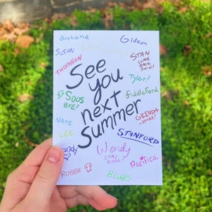 Könnte beinhalten: Eine weiße Karte mit der handgeschriebenen Nachricht "See you next Summer" in Schwarz, umgeben von farbenfrohen Namen und Phrasen. Die Karte wird vor einem Hintergrund aus grünem Laub gehalten.