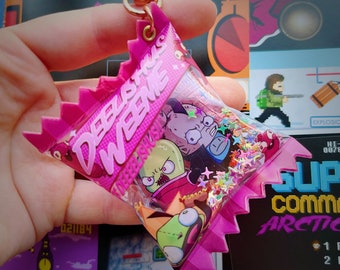 May include: Pink plastic keychain shaped like a candy bag with the text "DEBLISHUS WEEME" printed on the front. The bag contains glitter and small cartoon figures.