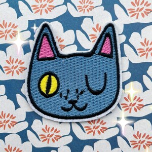 May include: A blue embroidered cat patch with a yellow eye and a pink ear. The cat has a winking expression.