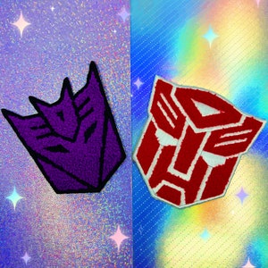 Może przedstawiać: Dwa haftowane naszywki z kultowymi logo Transformers. Lewa naszywka jest fioletowa i przedstawia symbol Autobotów, podczas gdy prawa naszywka jest czerwona i pokazuje symbol Decepticonów.