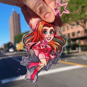 May include: A pink and red anime-style character with long red hair and a pink and white outfit. The character is holding a pink star and is surrounded by pink stars. The character is on a clear acrylic keychain.