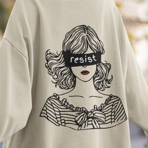 Può includere: Una felpa beige chiaro con una stampa grafica di una donna con una fascia nera sugli occhi che dice "resist".