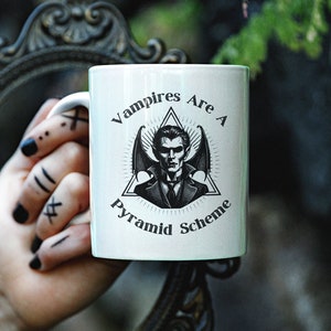 May include: White ceramic mug with a black and white illustration of a vampire with wings and the text "Vampires Are A Pyramid Scheme".