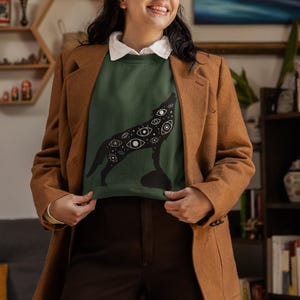 K&ouml;nnte beinhalten: Ein dunkelgr&uuml;nes Sweatshirt mit einer schwarzen Wolfsilhouette, die mit Augen bedeckt ist. Die Person tr&auml;gt einen braunen Blazer, ein wei&szlig;es Hemd mit Kragen und eine braune Hose. Der Hintergrund zeigt ein B&uuml;cherregal und Kunstwerke.