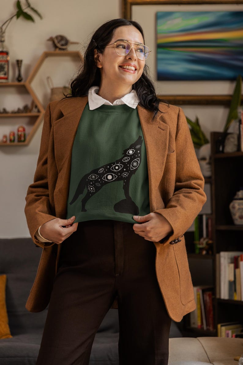 K&ouml;nnte beinhalten: Ein dunkelgr&uuml;nes Sweatshirt mit einer schwarzen Wolfsilhouette, die mit Augen bedeckt ist. Die Person tr&auml;gt einen braunen Blazer, ein wei&szlig;es Hemd mit Kragen und eine braune Hose. Der Hintergrund zeigt ein B&uuml;cherregal und Kunstwerke.