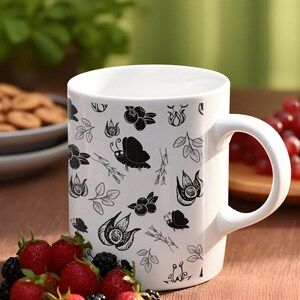 Puede incluir: Taza de cer&aacute;mica blanca con un patr&oacute;n bot&aacute;nico en blanco y negro con flores, hojas y mariposas. La taza est&aacute; llena de una bebida y est&aacute; rodeada de fresas y moras frescas. La taza tiene un asa curvada.