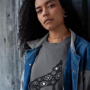 K&ouml;nnte beinhalten: Ein graues Sweatshirt mit einer schwarzen Illustration eines heulenden Wolfes, der mit Augen gef&uuml;llt ist. Die Person tr&auml;gt eine Jeansjacke &uuml;ber dem Sweatshirt. Der Hintergrund ist eine graue Wand.