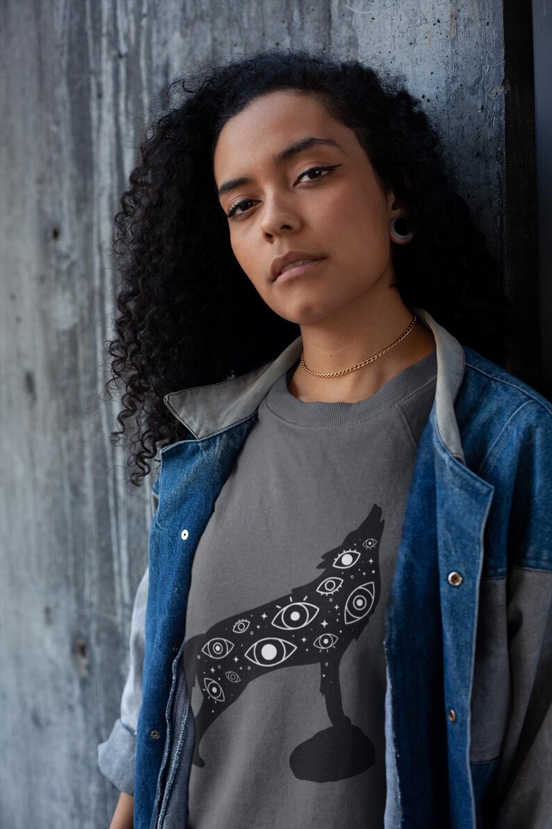 K&ouml;nnte beinhalten: Ein graues Sweatshirt mit einer schwarzen Illustration eines heulenden Wolfes, der mit Augen gef&uuml;llt ist. Die Person tr&auml;gt eine Jeansjacke &uuml;ber dem Sweatshirt. Der Hintergrund ist eine graue Wand.