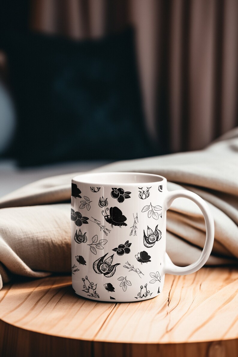Puede incluir: Taza de cer&aacute;mica blanca con un dise&ntilde;o floral y de mariposas en negro. La taza es cil&iacute;ndrica con un asa curvada. Se encuentra sobre una superficie de madera, con una tela de tono neutro y un fondo borroso.