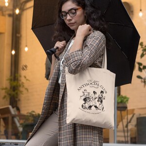 May include: A woman wearing a plaid jacket and beige pants is holding a black umbrella and a beige tote bag. The tote bag has a black and white illustration of women reading books and the text "The Ladies Antisocial Club Ignoring you, one book at a time."