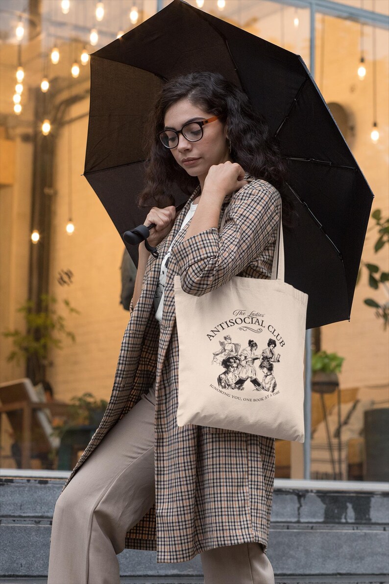 May include: A woman wearing a plaid jacket and beige pants is holding a black umbrella and a beige tote bag. The tote bag has a black and white illustration of women reading books and the text "The Ladies Antisocial Club Ignoring you, one book at a time."