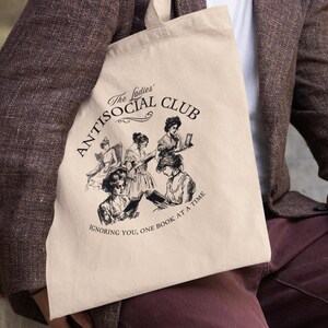 May include: A beige tote bag with a black graphic of women reading. The graphic says "The Ladies' Antisocial Club Ignoring You, One Book at a Time."