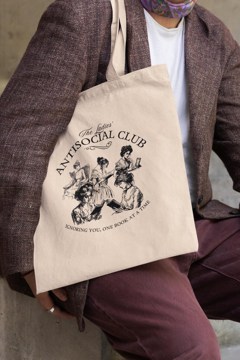 May include: A beige tote bag with a black graphic of women reading. The graphic says "The Ladies' Antisocial Club Ignoring You, One Book at a Time."