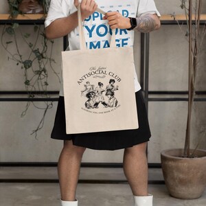 May include: A person is holding a beige tote bag with black handles. The bag has a black and white illustration of women reading books and the text "The ladies' Antisocial Club Honoring you, one book at a time."