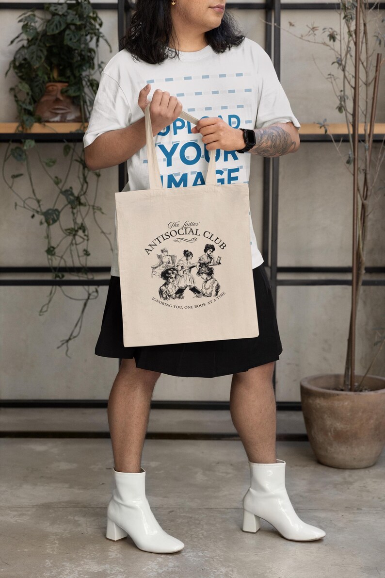 May include: A person is holding a beige tote bag with black handles. The bag has a black and white illustration of women reading books and the text "The ladies' Antisocial Club Honoring you, one book at a time."