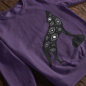 K&ouml;nnte beinhalten: Ein lila Sweatshirt mit einer schwarzen Silhouette eines heulenden Wolfes. Der Wolf ist mit wei&szlig;en Augen und Sternen gef&uuml;llt. Das Sweatshirt hat einen Rundhalsausschnitt und lange &Auml;rmel.
