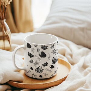 Puede incluir: Taza de cer&aacute;mica blanca con un patr&oacute;n bot&aacute;nico en blanco y negro con flores, hojas e insectos. La taza est&aacute; sobre una bandeja de madera, con un jarr&oacute;n de flores secas en el fondo. La taza tiene un asa curva.