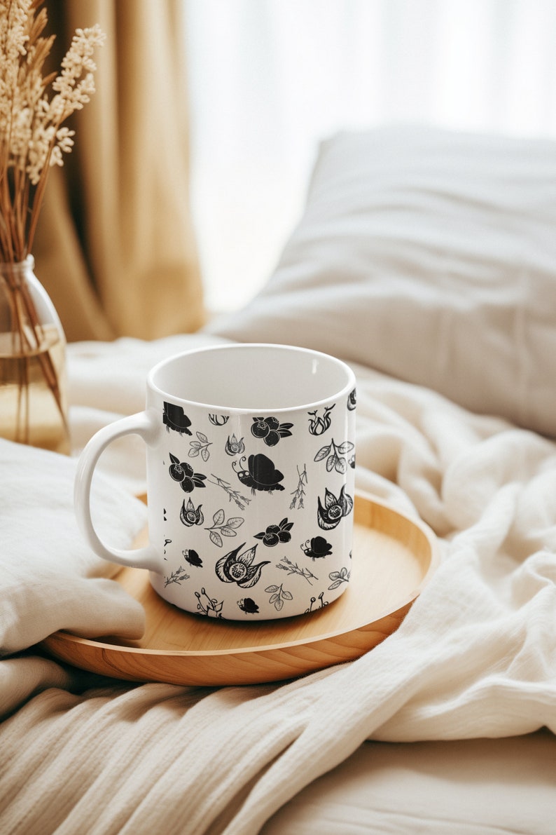 Puede incluir: Taza de cer&aacute;mica blanca con un patr&oacute;n bot&aacute;nico en blanco y negro con flores, hojas e insectos. La taza est&aacute; sobre una bandeja de madera, con un jarr&oacute;n de flores secas en el fondo. La taza tiene un asa curva.