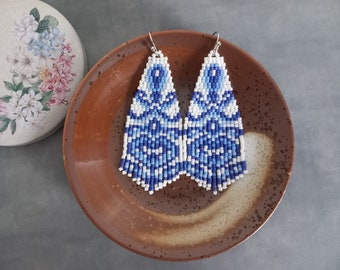 Op de afbeelding: Een paar kralen oorbellen in blauw en wit, gerangschikt op een bruine plaat. De oorbellen hebben een geometrisch patroon en een franje aan de onderkant. Een bloemversierde container is op de achtergrond.